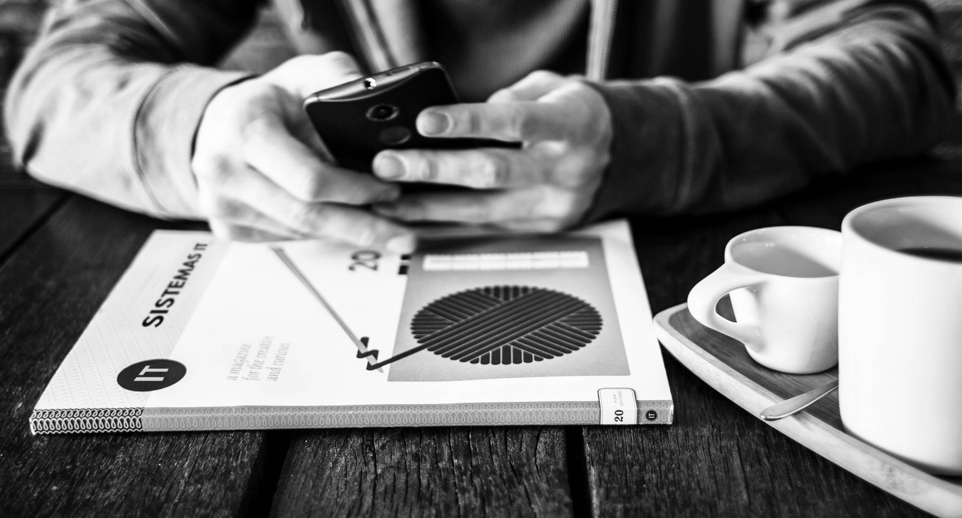 Manos de hombre sosteniendo un celular, apoyado sobre una mesa, junto a un libro de IT.